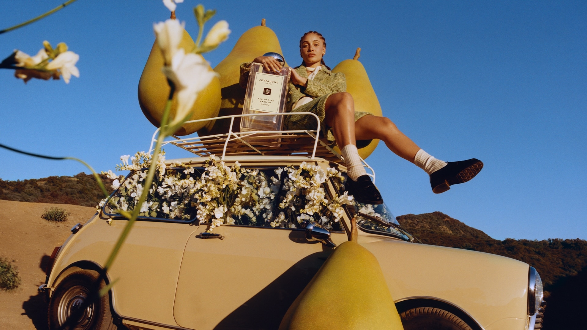 Adwoa Aboah with braided hair, biting into a pear with backdrop of three large pear props placed on top of a car roof rack.